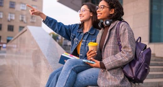 Deux jeunes femmes assises dehors, souriantes, l'une montrant du doigt au loin, tenant des cahiers et un café, avec un sac à dos.
